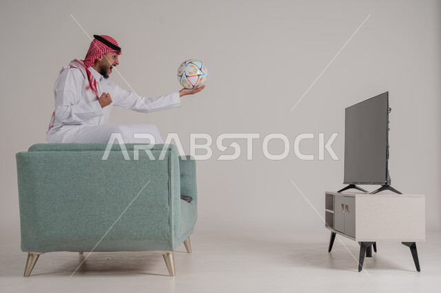 متابعة مباراة كرة القدم بتركيز وتمعن، بورتريه من الجانب لرجل عربي خليجي سعودي يجلس على كنبة مريحة امام التلفاز يمسك بالكرة ويشاهد لعب فريقه المفضل، رفع اليد بإيماءات الحماس والسعادة لإحراز هدف، تنمية شغف الاهتمامات الرياضية، الجسم بالطول الكامل، خلفية رمادية