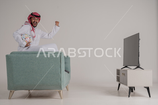 متابعة مباراة كرة القدم بتركيز وتمعن، بورتريه من الجانب لرجل عربي خليجي سعودي يجلس على كنبة مريحة امام التلفاز يمسك بالكرة ويشاهد لعب فريقه المفضل، رفع اليد بإيماءات الحماس والسعادة لإحراز هدف، تنمية شغف الاهتمامات الرياضية، الجسم بالطول الكامل، خلفية رمادية