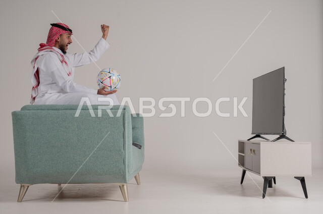 Watching a football match with concentration and contemplation, side portrait of a Saudi Arabian Gulf man sitting on a comfortable sofa in front of the TV holding the ball and watching his favorite team play, raising hand with gestures of enthusiasm and happiness for scoring a goal, developing passion for sports interests, full body, gray background