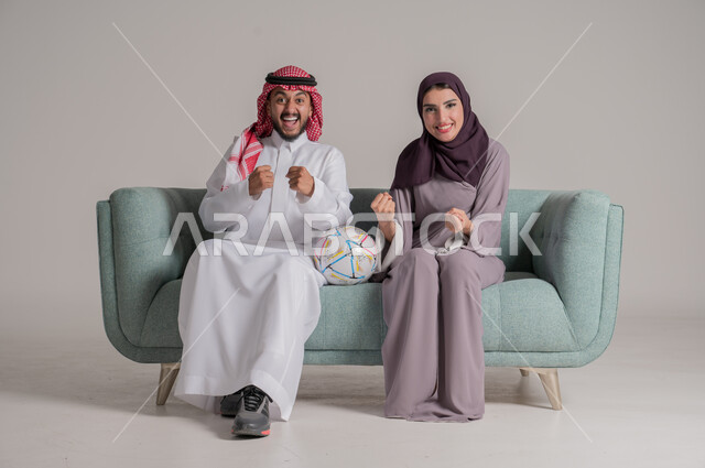 Enjoying cheering for your favorite team and watching league matches at home, side portrait of a Saudi Arabian Gulf couple wearing traditional clothes sitting on a comfortable sofa in the living room watching the match together with gestures of enthusiasm and integration, full body, gray background
