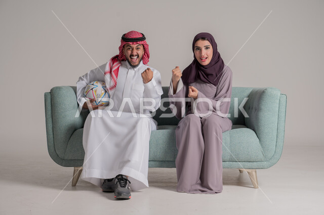 الاستمتاع بتشجيع الفريق المفضل ومتابعة مباريات الدوري في المنزل، بورتريه من الجانب لزوجين عربيين خليجيين سعوديين يرتديان الملابس التقليدية يجلسان على كنبة مريحة في غرفة المعيشة ويشاهدان المباراة سويا بإيماءات الحماس والاندماج، الجسم بالطول الكامل، خلفية رمادية