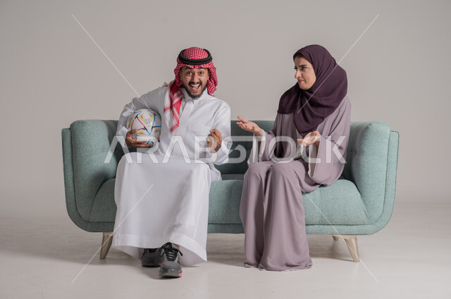 الاستمتاع بتشجيع الفريق المفضل ومتابعة مباريات الدوري في المنزل، بورتريه من الجانب لزوجين عربيين خليجيين سعوديين يرتديان الملابس التقليدية يجلسان على كنبة مريحة في غرفة المعيشة ويشاهدان المباراة سويا بإيماءات الحماس والاندماج، الجسم بالطول الكامل، خلفية رمادية