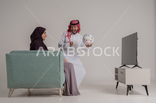 تشجيع الفريق المفضل ومتابعة المباراة في المنزل، بورتريه من الجانب لزوجين عربيين خليجيين سعوديين يجلسان على كنبة مريحة أمام التلفاز، رجل يرتدي الشماغ والثوب التقليدي يمسك الكرة ويقفز بإيماءات الفرح والحماس بإحراز هدف الفوز، الجسم بالطول الكامل، خلفية رمادية