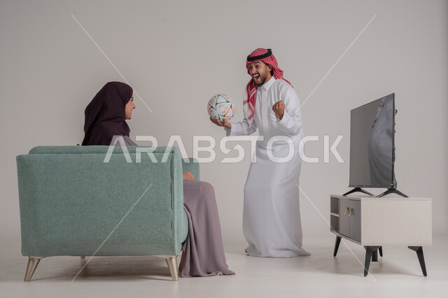 تشجيع الفريق المفضل ومتابعة المباراة في المنزل، بورتريه من الجانب لزوجين عربيين خليجيين سعوديين يجلسان على كنبة مريحة أمام التلفاز، رجل يرتدي الشماغ والثوب التقليدي يمسك الكرة ويقفز بإيماءات الفرح والحماس بإحراز هدف الفوز، الجسم بالطول الكامل، خلفية رمادية