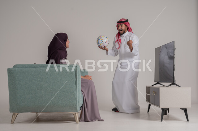 تشجيع الفريق المفضل ومتابعة المباراة في المنزل، بورتريه من الجانب لزوجين عربيين خليجيين سعوديين يجلسان على كنبة مريحة أمام التلفاز، رجل يرتدي الشماغ والثوب التقليدي يمسك الكرة ويقفز بإيماءات الفرح والحماس بإحراز هدف الفوز، الجسم بالطول الكامل، خلفية رمادية