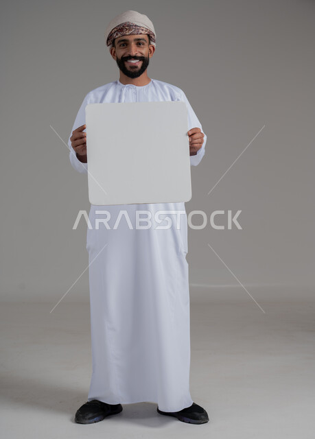 الإشارة إلى لوحة بيضاء مربعة فارغة، إعلانات وعروض تسويقية، استخدام لوحات توضيحية ووسائل تعليمية، بورتريه لشاب عربي خليجي عماني مبتسم يرتدي يرتدي الدشداشة والعمامة يحمل بيده موك اب ابيض تظهر عليه علامات السعادة والسرور, صورة الجسم بالكامل، خلفية بيضاء