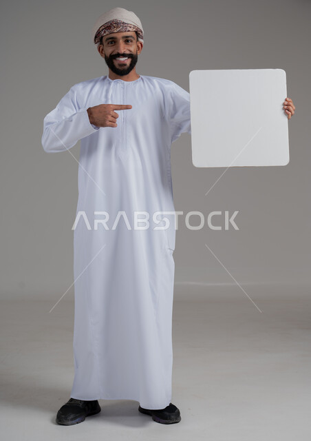 الإشارة إلى لوحة بيضاء مربعة فارغة، إعلانات وعروض تسويقية، استخدام لوحات توضيحية ووسائل تعليمية، بورتريه لشاب عربي خليجي عماني مبتسم يرتدي يرتدي الدشداشة والعمامة يحمل بيده موك اب ابيض تظهر عليه علامات السعادة والسرور, صورة الجسم بالكامل، خلفية بيضاء