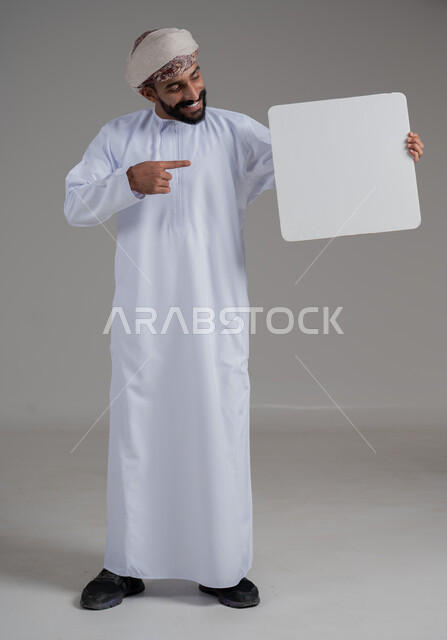 الإشارة إلى لوحة بيضاء مربعة فارغة، إعلانات وعروض تسويقية، استخدام لوحات توضيحية ووسائل تعليمية، بورتريه لشاب عربي خليجي عماني مبتسم يرتدي يرتدي الدشداشة والعمامة يحمل بيده موك اب ابيض تظهر عليه علامات السعادة والسرور, صورة الجسم بالكامل، خلفية بيضاء