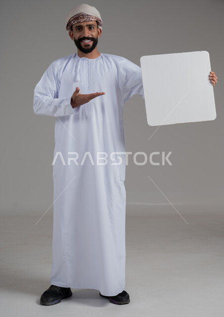 Pointing to a blank square white board, advertisements and marketing offers, using illustration boards and educational tools, portrait of a smiling young Arab Gulf Omani man wearing a dishdasha and turban holding a white mockup showing signs of happiness and pleasure, full body image, white background