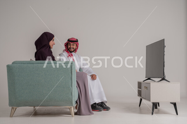 تقوية روابط المحبة والمودة، الاستمتاع بقضاء أوقات الفراغ مع الأسرة في المنزل، بورتريه من الجانب لزوجين عربيين خليجيين سعوديين يرتديان الملابس التقليدية يجلسان على كنبة مريحة في غرفة المعيشة ويشاهدان التلفاز في جو يسوده الهدوء، الجسم بالطول الكامل، خلفية رمادية