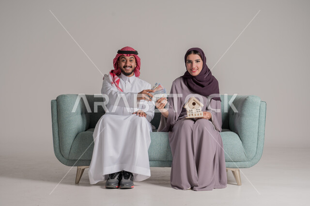 بناء أسرة ناجحة متفاهمة ومتماسكة، الإمساك بمجسم صغير للبيت بإيماءات السعادة، بورتريه لزوجين عربيين خليجيين سعوديين يرتديان الملابس التقليدية يجلسان على كنبة مريحة في غرفة المعيشة ويضعان الخطط الاقتصادية لإدارة مصاريف المنزل، الجسم بالطول الكامل، خلفية رمادية