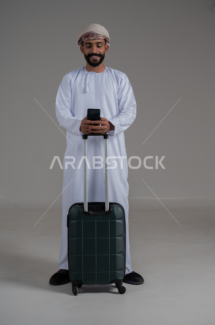 الاستعداد لقضاء رحلة صيفية ممتعة, الإتفاق على موعد الانطلاق مع الأصدقاء، مفهوم السياحة في إجازة الصيف، القيام بالجولات الترفيهية، بورتريه لشاب عربي عماني يرتدي الدشداشة والعمامة و يحمل بيده هاتف محمول وبجانبه حقيبة الامتعة، الجسم بالطول الكامل, خلفية بيضاء