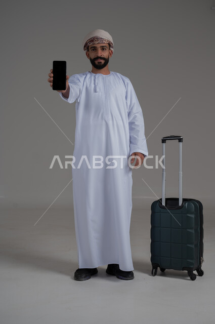 الاستعداد لقضاء رحلة صيفية ممتعة، مفهوم السياحة في إجازة الصيف، القيام بالجولات الترفيهية، بورتريه لشاب عربي عماني يرتدي الدشداشة والعمامة يقوم بعرض شاشة سوداء فارغة وبجانبه حقيبة الامتعة، الجسم بالطول الكامل, خلفية بيضاء
