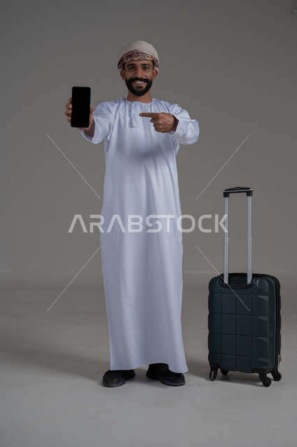 الاستعداد لقضاء رحلة صيفية ممتعة، مفهوم السياحة في إجازة الصيف، القيام بالجولات الترفيهية، بورتريه لشاب عربي عماني يرتدي الدشداشة والعمامة يقوم بعرض شاشة سوداء فارغة وبجانبه حقيبة الامتعة، الجسم بالطول الكامل, خلفية بيضاء