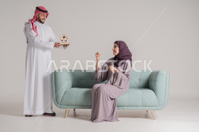 بناء أسرة ناجحة متفاهمة ومتماسكة، الإمساك بمجسم صغير للبيت والإشارة إليه بإيماءات السعادة والأمل، بورتريه لزوجين عربيين خليجيين سعوديين يرتديان الملابس التقليدية يقضيان وقتهما في غرفة المعيشة في جو من المحبة والمودة العائلية، الجسم بالطول الكامل، خلفية رمادية