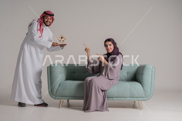 بناء أسرة ناجحة متفاهمة ومتماسكة، الإمساك بمجسم صغير للبيت والإشارة إليه بإيماءات السعادة والأمل، بورتريه لزوجين عربيين خليجيين سعوديين يرتديان الملابس التقليدية يقضيان وقتهما في غرفة المعيشة في جو من المحبة والمودة العائلية، الجسم بالطول الكامل، خلفية رمادية