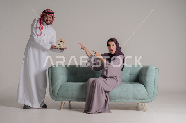 Building a successful, understanding and cohesive family, holding a small model of the house and pointing to it with gestures of happiness and hope, portrait of a Saudi Arabian Gulf couple wearing traditional clothes spending their time in the living room in an atmosphere of love and family affection, full length body, gray background