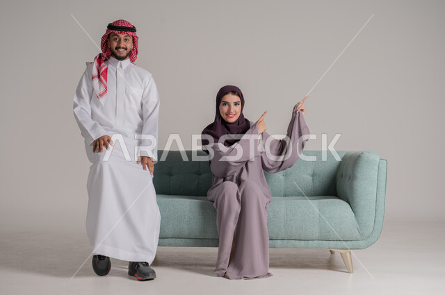 تقوية روابط المحبة والمودة، الاستمتاع بقضاء أوقات الفراغ مع الأسرة في المنزل، بورتريه لزوجين عربيين خليجيين سعوديين يرتديان الملابس التقليدية يجلسان على كنبة مريحة في غرفة المعيشة ينظران للكاميرا بإيماءات السرور والسعادة، الجسم بالطول الكامل، خلفية رمادية