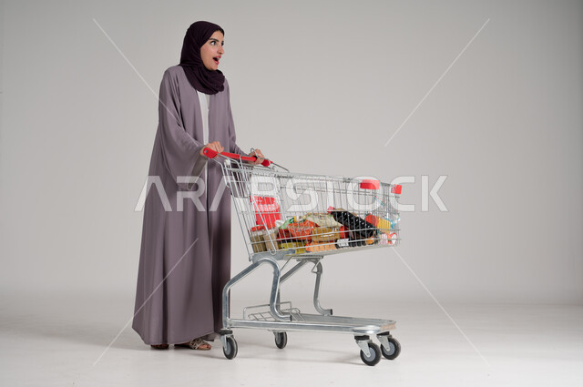 الاستمتاع بالتسوق في المولات والمراكز التجارية، شراء المستلزمات والحاجيات الضرورية، الاستفادة من الخصومات على المنتجات، بورتريه لامرأة عربية خليجية سعودية محجبة ترتدي العباءة التقليدية وتجر عربة التسوق أمامها بإيماءات السعادة، الجسم بالطول الكامل، خلفية رمادية