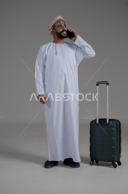 الاتفاق مع الاصدقاء على موعد الانطلاق, الاستعداد لقضاء رحلة صيفية ممتعة، مفهوم السياحة في إجازة الصيف، القيام بالجولات الترفيهية، بورتريه لشاب عربي عماني يرتدي الدشداشة والعمامة يقوم بإجراء مكالمة هاتفية وبجانبه حقيبة الامتعة، الجسم بالطول الكامل, خلفية بيضاء