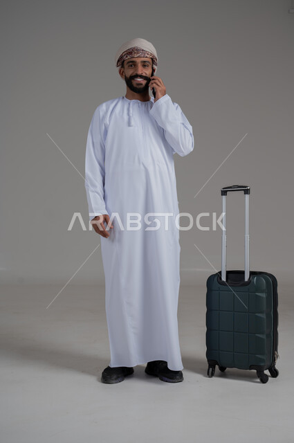 الاتفاق مع الاصدقاء على موعد الانطلاق, الاستعداد لقضاء رحلة صيفية ممتعة، مفهوم السياحة في إجازة الصيف، القيام بالجولات الترفيهية، بورتريه لشاب عربي عماني يرتدي الدشداشة والعمامة يقوم بإجراء مكالمة هاتفية وبجانبه حقيبة الامتعة، الجسم بالطول الكامل, خلفية بيضاء