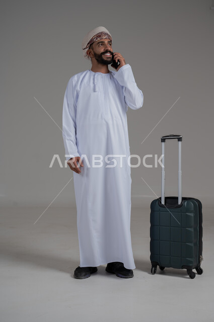 الاتفاق مع الاصدقاء على موعد الانطلاق, الاستعداد لقضاء رحلة صيفية ممتعة، مفهوم السياحة في إجازة الصيف، القيام بالجولات الترفيهية، بورتريه لشاب عربي عماني يرتدي الدشداشة والعمامة يقوم بإجراء مكالمة هاتفية وبجانبه حقيبة الامتعة، الجسم بالطول الكامل, خلفية بيضاء