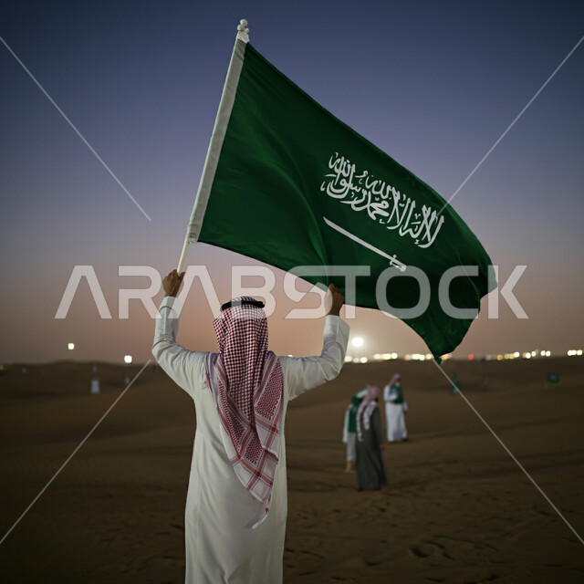 إستذكار التاريخ والهوية الوطنية، صورة من الخلف لرجل عربي خليجي سعودي يرتدي الشماغ والثوب ويحمل علم السعودية، العيد الوطني 8 ربيع الأول، يوم العلم السعودي 11 مارس، غروب الشمس في الصحراء