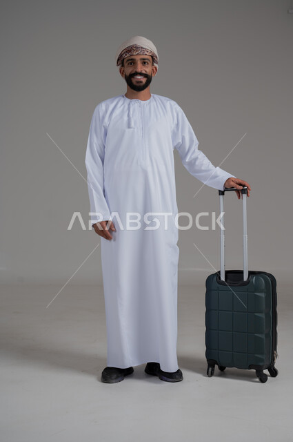 الاستعداد لقضاء رحلة صيفية ممتعة، مفهوم السياحة في إجازة الصيف، القيام بالجولات الترفيهية، بورتريه لشاب عربي عماني يرتدي الدشداشة والعمامة ينظر الى الكاميرا بتعابير السعادة والسرور وبجانبه حقيبة الامتعة، الجسم بالطول الكامل, خلفية بيضاء