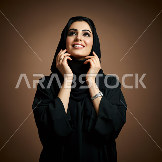 الاعتناء بالمظهر الخارجي والشياكة والأناقة, تعابير السعادة والسرور ، بورتريه مقرب لإمرأة عربية خليجية سعودية محجبة مبتسمة ترتدي العباءة السوداء تنظر إلى مكان ما في الأعلى بإيماءات الثقة بالنفس والفرح، مفهوم الأنوثة والنعومة لدى النساء السعوديات، خلفية بنية