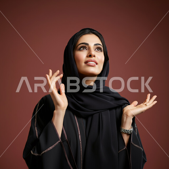 النظر والتأمل بإيماءات السعادة والسرور، مفهوم النعومة والأنوثة، الاناقة والاهتمام بالمظهر الخارجي، بورتريه مقرب لإمرأة عربية خليجية سعودية محجبة مبتسمة ترتدي العباءة السوداء تنظر إلى الأعلى بتعابير الإنبهار والإعجاب بشيء ما، خلفية بنية