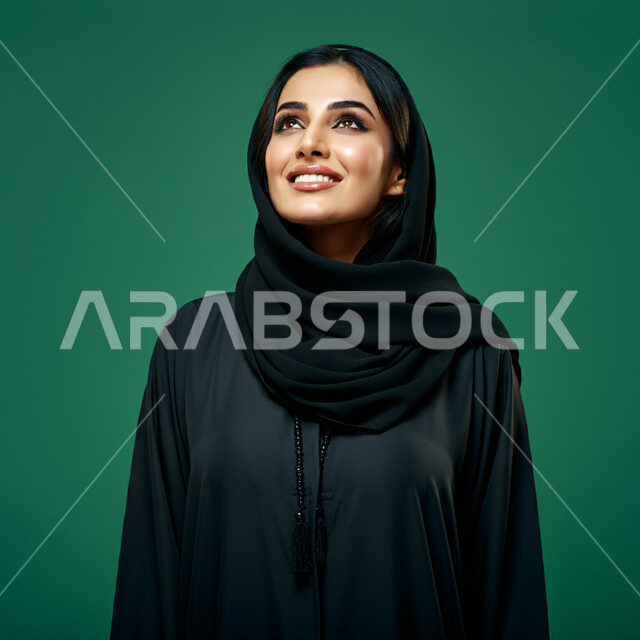 مفهوم الأنوثة والنعومة، بورتريه مقرب لامرأة عربية خليجية سعودية محجبة مبتسمة ترتدي العباءة السوداء تنظر إلى الأعلى  بإيماءات الثقة بالنفس والفرح، الاهتمام بالأناقة والمظهر الخارجي، تعابير السرور والسعادة، خلفية خضراء، كروما