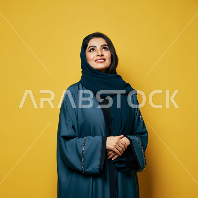 النظر والتأمل بإيماءات السعادة والسرور، مفهوم الأنوثة والنعومة، بورتريه مقرب لامرأة عربية خليجية سعودية محجبة مبتسمة ترتدي العباءة الزرقاء تنظر إلى الأعلى  بتعابير الثقة بالنفس والفرح، الأعتناء بالأناقة والمظهر الخارجي، خلفية صفراء