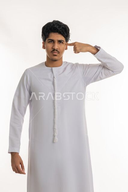 الإصرار على الرأي و فعل ما يرغب, مفهوم التفكير والتحليل و الذكاء والفطنة, بورتريه لشاب مراهق عربي خليجي إماراتي يرتدي الكندورة يقف باستقامة و يشير بإصبع السبابة إلى رأسه بتعابير العناد, خلفية بيضاء