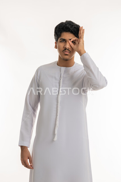 التركيز والنظر إلى شيء محدد، محاولة تقريب الأشياء البعيدة, مفهوم التخطيط و الرؤية المستقبلية، بورتريه لشاب عربي خليجي اماراتي يرتدي الكندورة يضع يده على عينيه بشكل عدسة بإيماءات البحث والاستكشاف، خلفية بيضاء