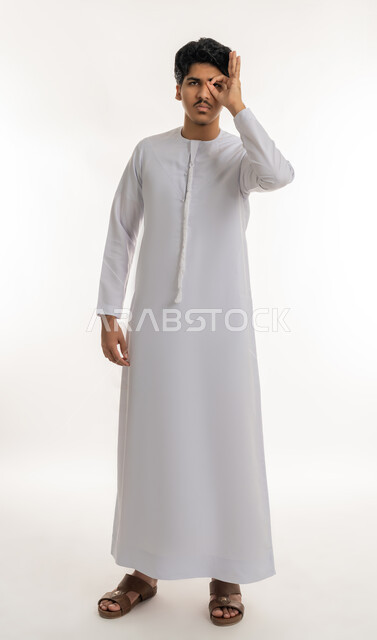 محاولة تقريب الأشياء البعيدة, مفهوم التخطيط و الرؤية المستقبلية، بورتريه لشاب عربي خليجي اماراتي يرتدي الكندورة يضع يده على عينيه بشكل عدسة بإيماءات البحث والاستكشاف، التركيز والنظر إلى شيء محدد، الطول الكامل للجسم، خلفية بيضاء