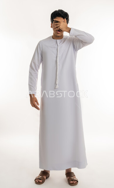 تذكر شيء ما بشكل مفاجئ، بورتريه لشاب عربي خليجي إماراتي يرتدي الكندورة يغطي عينيه بيده، ايماءات الاستغراب والمفاجأة، مراقبة شيء ما، أخذ نظرة خاطفة على موقف محدد، تعابير الخجل والحياء، الطول الكامل للجسم، خلفية بيضاء