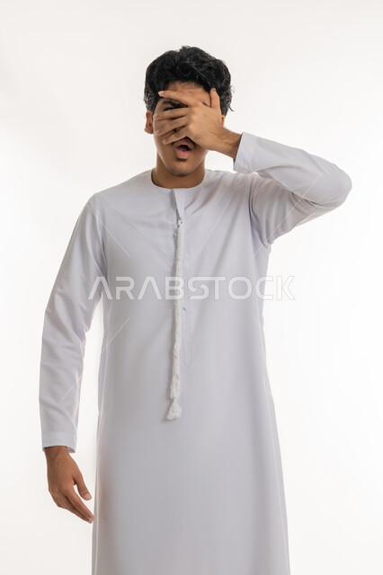 تعابير الخجل والحياء، تذكر شيء ما بشكل مفاجئ، بورتريه لشاب عربي خليجي إماراتي يرتدي الكندورة يغطي عينيه بيده، ايماءات الاستغراب والمفاجأة، أخذ نظرة خاطفة على موقف محدد، خلفية بيضاء