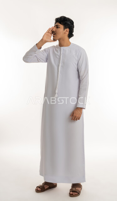 التحدث بصوت منخفض، مفهوم المناداة بصوت مرتفع، بورتريه لشاب عربي خليجي إماراتي يرتدي الكندورة يقوم بالنداء خلف يده، كتمان وحفظ الاسرار، مشاركة سر ما، الهمس والهدوء اثناء الكلام، الطول الكامل للجسم، خلفية بيضاء