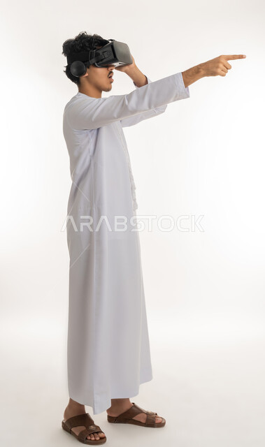 رؤية مستقبلية بعيدة، بورتريه لشاب عربي خليجي إماراتي يرتدي الكندورة ونظارات الواقع الافترضي ثلاثية الابعاد يشير بإصبع السبابة الى مكان بعيد، محاكاة العالم الخيالي، أجهزة تكنولوجية وتقنيات حديثة، الطول الكامل للجسم، خلفية بيضاء