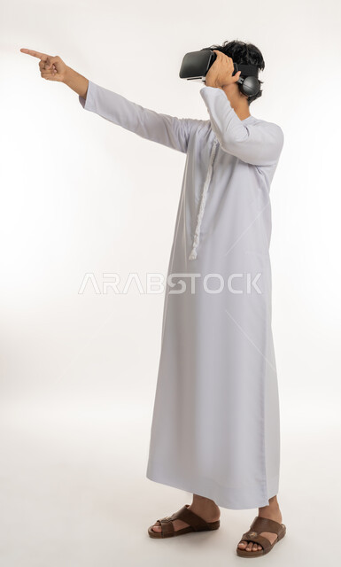 Simulating the imaginary world, technological devices and modern technologies, distant future vision, portrait of an Arab young man Emirati Gulf man wearing kandura and 3D virtual reality glasses pointing index finger to distant place, full body length, white background