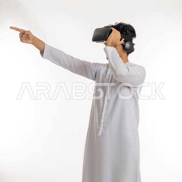 تعابير الاندماج والمتعة, محاكاة العالم الخيالي، أجهزة تكنولوجية وتقنيات حديثة، رؤية مستقبلية بعيدة، بورتريه لشاب عربي خليجي إماراتي يرتدي الكندورة ونظارات الواقع الافترضي ثلاثية الابعاد يشير بإصبع السبابة الى مكان بعيد، خلفية بيضاء