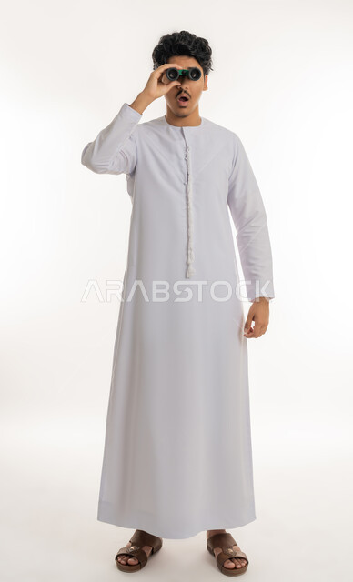 Searching for something and expanding the field of vision using binoculars, concept of exploration and adventure, future visions among young people, portrait of a young Arab Gulf Emirati man wearing a kandura looking through binoculars, gestures of surprise and astonishment, full body length, white background