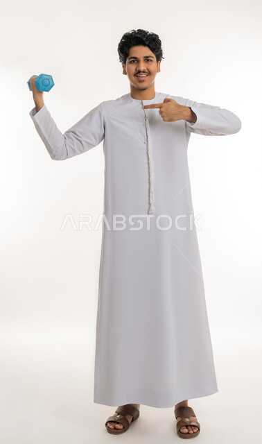Physical activity to maintain and physical fitness, portrait of a plump young Arab Gulf Emirati man wearing a kandura doing dumbbell exercises, strengthening and building body muscles, pointing with the index finger on heavy iron weights, full body length, white background