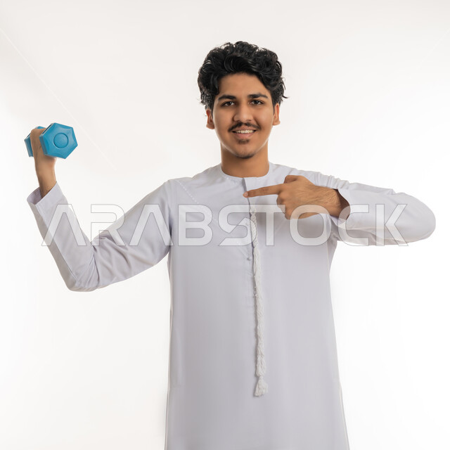 الاشارة بإصبع السبابة على الأوزان الحديدية الثقيلة، اتباع نظام صحي، نشاط بدني للمحافظة على و اللياقة الجسدية، بورتريه لشاب عربي خليجي اماراتي ممتلئ يرتدي الكندورة يقوم بممارسة التمارين الرياضية بالدمبل، تقوية عضلات الجسم و بنائها، خلفية بيضاء