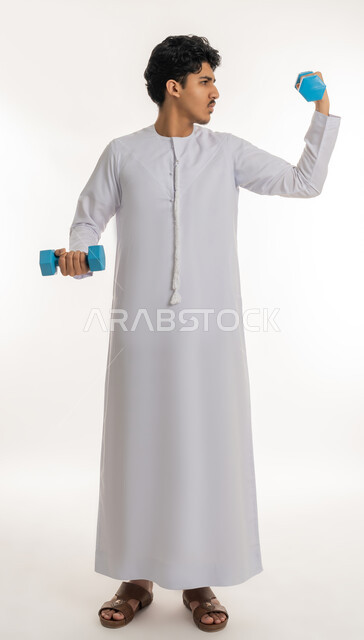 تقوية عضلات الجسم و بنائها، رفع الأوزان الحديدية الثقيلة، اتباع نظام صحي، نشاط بدني للمحافظة على الصحة و اللياقة الجسدية، بورتريه لشاب عربي خليجي اماراتي ممتلئ يرتدي الكندورة يقوم بممارسة التمارين الرياضية بالدمبل، الطول الكامل للجسم، خلفية بيضاء