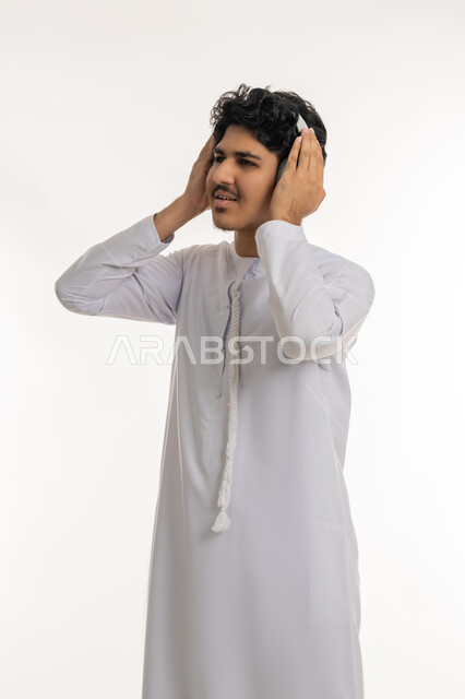 الاندماج مع الأنغام الموسيقية, قضاء أوقات ممتعة بالإستماع للأغاني والمواهب الغنائية، مفهوم الإصغاء باسخدام الهيدفون، بورتريه لشاب عربي خليجي إماراتي يرتدي الكندورة يضع سماعات الرأس بإيماءات الفرح والسرور، خلفية بيضاء