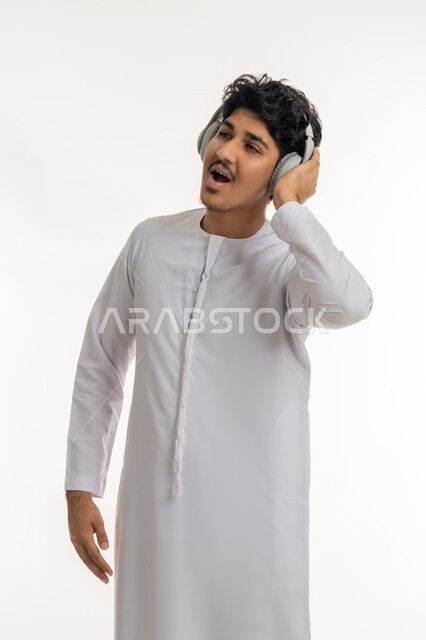 مفهوم الإصغاء باسخدام الهيدفون، بورتريه لشاب عربي خليجي إماراتي يرتدي الكندورة يضع سماعات الرأس بإيماءات الفرح والسرور، التفاعل والاندماج مع الأنغام الموسيقية, الاستماع للأغاني والمواهب الغنائية، خلفية بيضاء