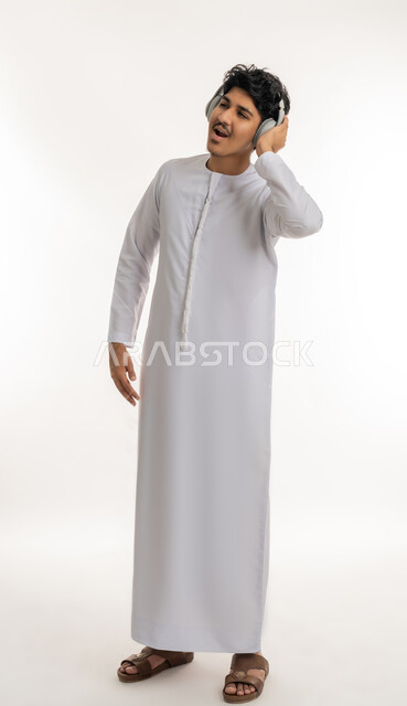 التفاعل والاندماج مع الأنغام الموسيقية, الاستماع للأغاني والمواهب الغنائية، مفهوم الإصغاء باسخدام الهيدفون، بورتريه لشاب عربي خليجي إماراتي يرتدي الكندورة يضع سماعات الرأس بإيماءات الفرح والسرور، الطول الكامل للجسم، خلفية بيضاء