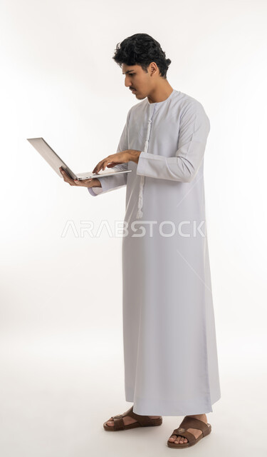 انجاز الاعمال المطلوبة، دمج التكنولوجيا بالحياة العملية، بورتريه لشاب عربي خليجي إماراتي يرتدي الكندورة يقف ويمسك بيده الحاسب المحمول، تصفح مواقع التواصل الإجتماعي، ايماءات التركيز والاندماج، مشاهدة الأفلام والمسلسلات، الطول الكامل للجسم، خلفية بيضاء