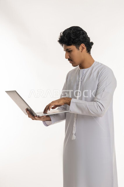 ايماءات التركيز والاندماج، مشاهدة الأفلام والمسلسلات، انجاز الاعمال المطلوبة، دمج التكنولوجيا بالحياة العملية، بورتريه لشاب عربي خليجي إماراتي يرتدي الكندورة يقف ويمسك بيده الحاسب المحمول، تصفح مواقع التواصل الإجتماعي، خلفية بيضاء